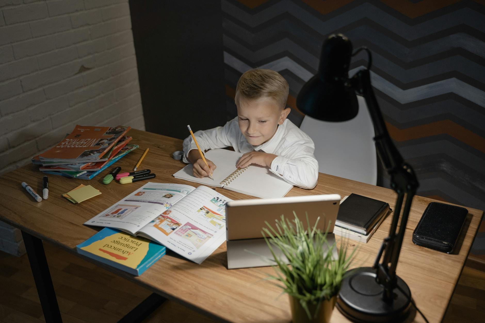 Chłopiec uczy się przy drewnianym biurku z tabletem i książkami pod lampką